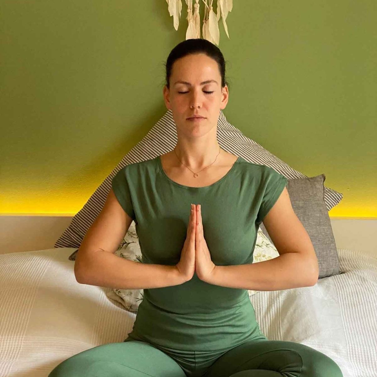 Yogini trägt einen grünen Jumpsuit, sitzt auf dem Bett und meditiert