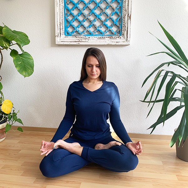Die Haltung bei der Achtsamkeitsmeditation Yogini sitzt im Lotussitz und meditiert, sie trägt des Yoga Longsleeve Matangi und die Hose Tara in Marine