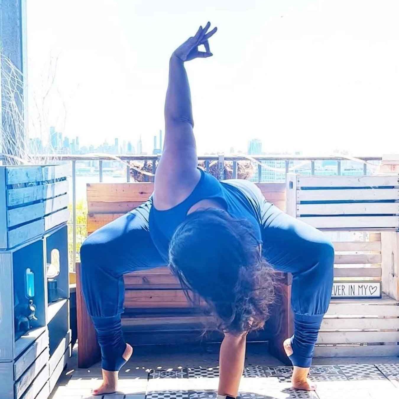 Frau steht in einer Yogapose auf dem Boden eines Balkons und trägt einen blauen Overall mit Stulpen. Sie stützt sich vornüber gebeugt mit einer Hand ab, die andere hält sie über sich im Chin Mudra.