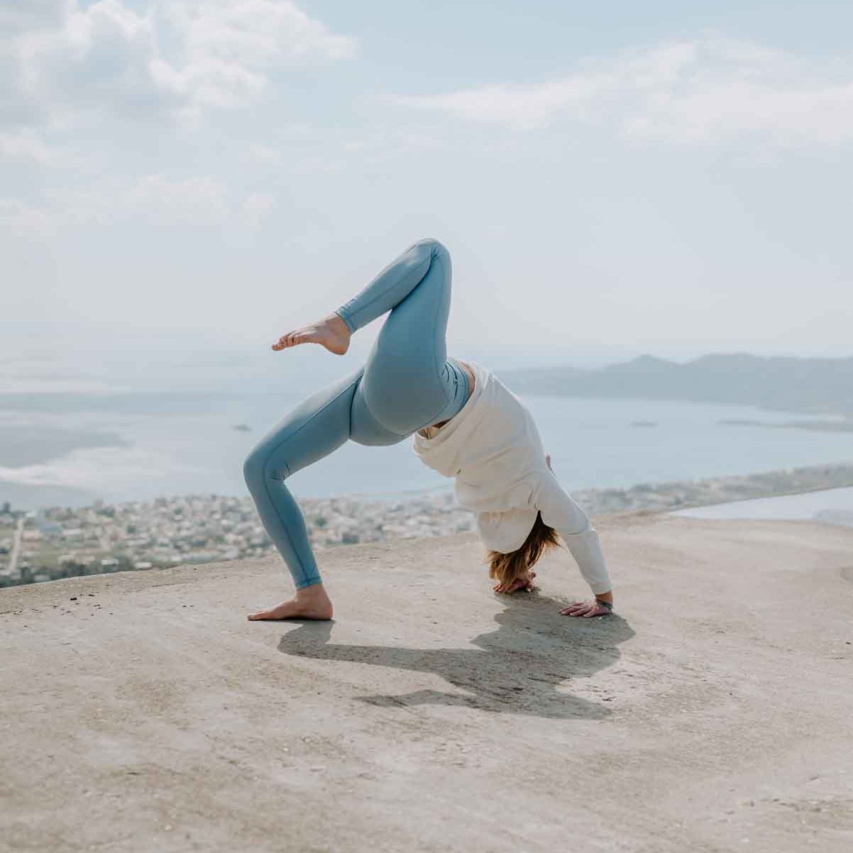 Frau macht draußen auf einem Plateau vor einer Küstenstadt Yoga in einem Outfit von chakrana