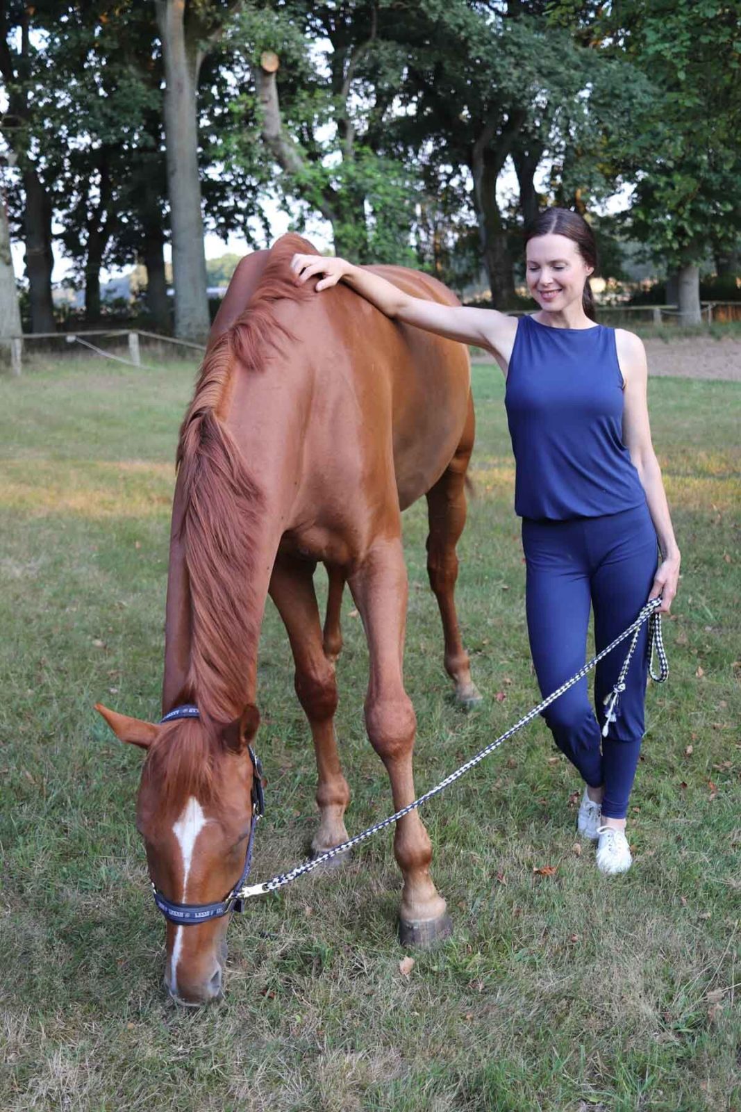 Eine Frau steht in einem Yoga-Overall auf einem Rasen neben einem braunen Pferd und hält es zärtlich an Führstrick und Schulter.